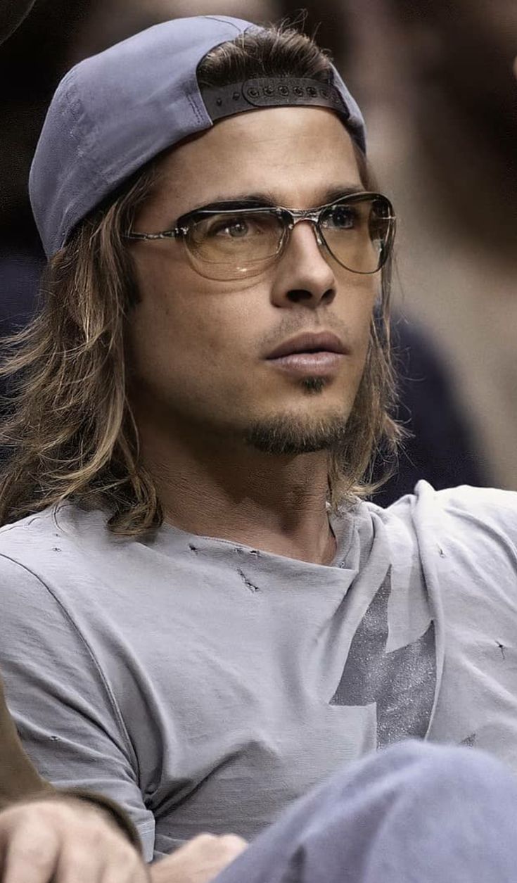 a man with long hair wearing glasses and a baseball cap sitting in the stands at a game