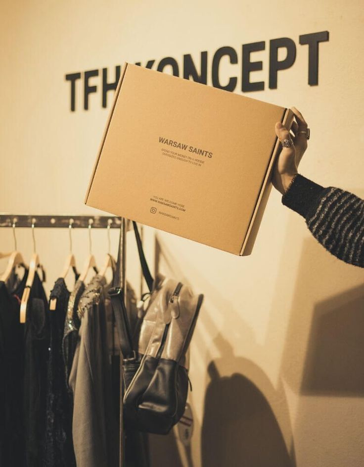 a person holding up a box in front of a rack with clothes on hangers