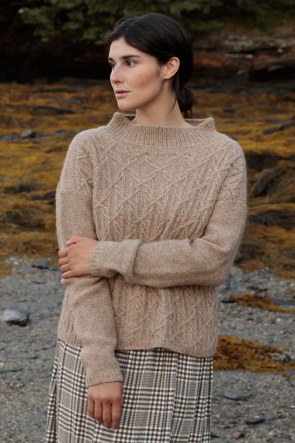 a woman standing in front of some rocks wearing a brown sweater and plaid skirt with her arms crossed