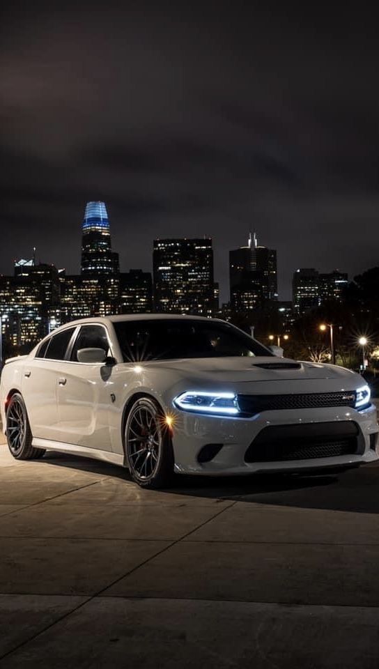 a white dodge charger parked in front of a cityscape at night with the lights on