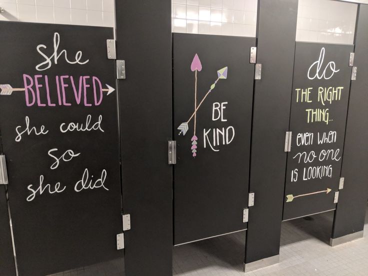 three black stalls with writing on them in a public bathroom stall, one has a pink flower and the other says she believed