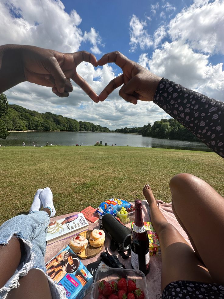 two people making a heart shape with their hands over a picnic table filled with food