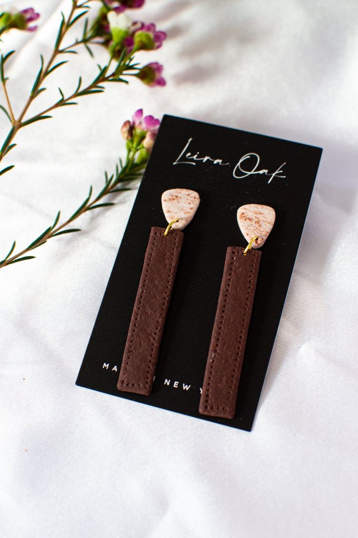 a pair of brown leather earrings sitting on top of a white sheet next to purple flowers