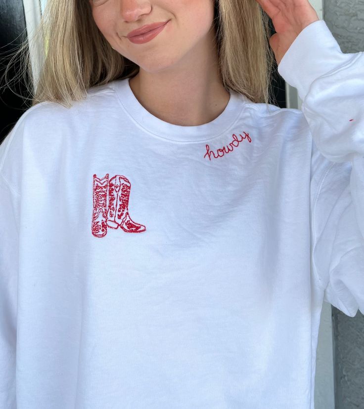 Introducing our "Howdy Embroidered Crewneck" - a fusion of comfort and Western charm that will effortlessly elevate your wardrobe. This exquisite white crewneck is not just an article of clothing; it's a statement piece that embraces the spirit of the frontier in a contemporary fashion. At the heart of this design lies the word "Howdy," hand embroidered in vibrant red cursive around the neckline. This subtle yet bold touch adds a warm and welcoming vibe, making it a perfect choice for casual outings and gatherings with friends. The real showstopper, however, is the finely detailed red cowboy boots embroidery adorning the right side of the chest. These intricate boots pay homage to the timeless allure of the Wild West, offering a tasteful nod to Western heritage while keeping the design mod Oversized Embroidered White Top, Oversized White Embroidered Top, White Custom Embroidery Relaxed Fit Tops, White Tops With Custom Embroidery Relaxed Fit, White Relaxed Fit Top With Custom Embroidery, White Embroidered Crew Top, White Embroidered Sweatshirt For Fall, White Tops With Embroidered Text For Fall, Cowboy Embroidery