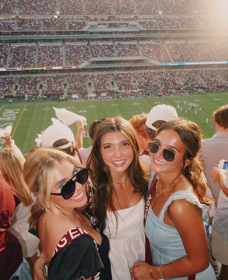 three girls are posing for the camera in front of a large crowd at a football game