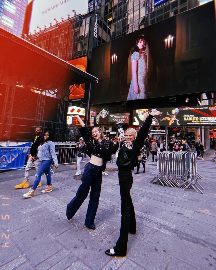 two women standing in front of a large screen on the side of a building with their arms up