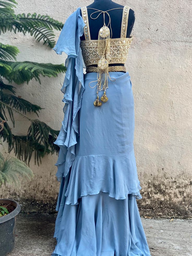 A two-piece blue ruffle saree set from the Priti Sahni collection. This beautiful blue georgette draped ruffle saree is paired with our classic mirror and pearl hand embroidered bustier blouse. The blouse has mirror-sequin tassels tie-up at the back. The outfit is completed with a heavy embroidered pearl belt.
Belt Included. Festive Draped Choli With Ruffles, Festive Draped Ruffle Choli, Festive Draped Ruffles Choli, Festive Draped Ruffled Choli, Designer Semi-stitched Ruffled Blouse Piece, Designer Ruffled Blouse Piece For Diwali, Designer Bollywood Style Ruffled Sharara, Designer Bollywood Style Sharara With Ruffles, Traditional Draped Sets With Ruffles