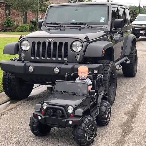 a baby sitting on top of a toy jeep in front of a black jeep truck