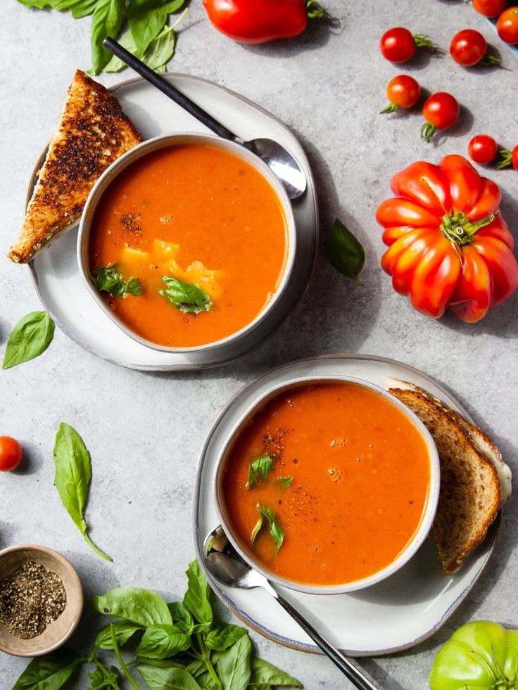 two bowls of tomato soup on a table with bread, tomatoes and basil around them