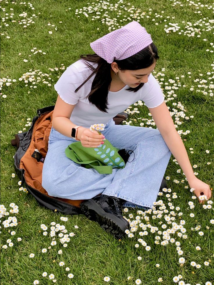 a woman sitting in the grass with a bag and some flowers on the ground next to her