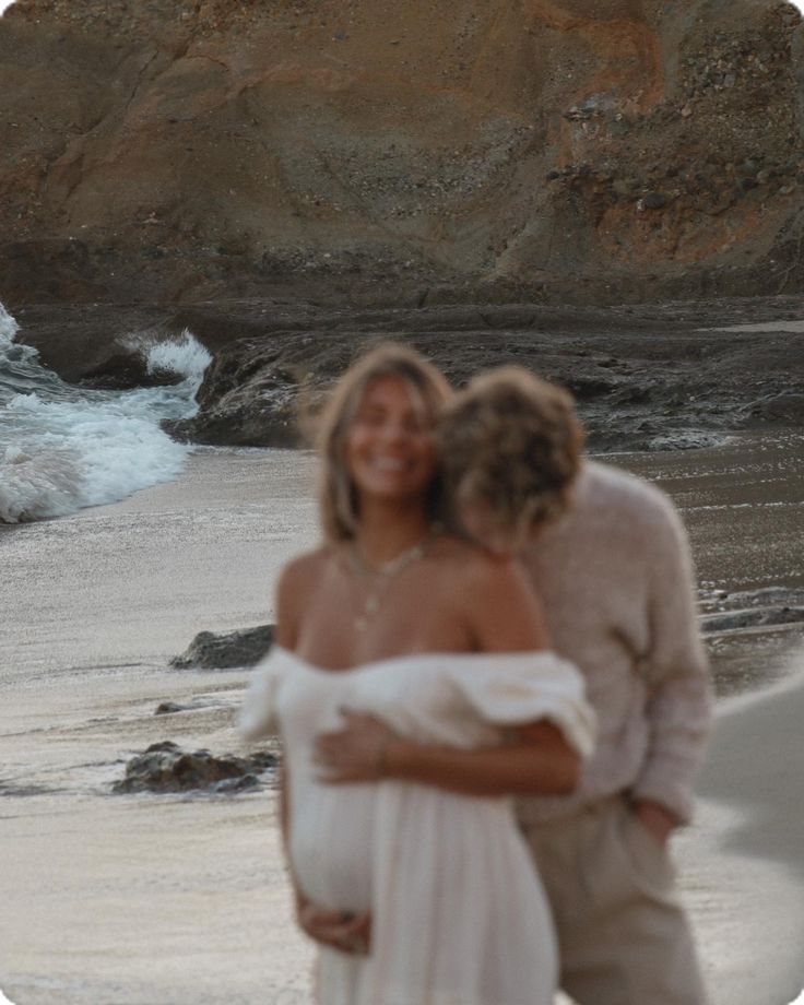 a man and woman hugging on the beach next to the ocean with waves coming in