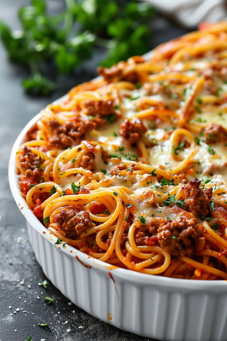 a casserole dish filled with spaghetti and meat sauce on a gray surface next to parsley