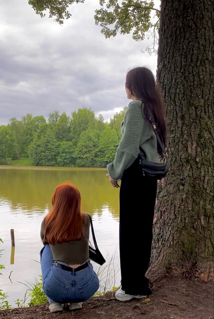 two women sitting on the ground next to a tree looking out over a body of water