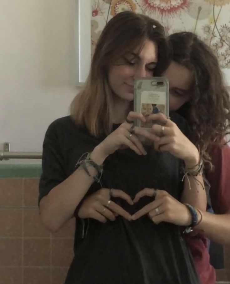 two young women standing in front of a mirror looking at their cell phones and making a heart shape with their hands