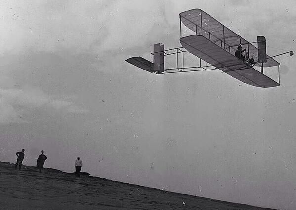 an old photo with people standing on top of a hill watching a plane fly overhead