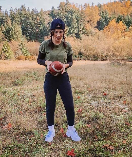 a woman holding a football in her right hand while standing in a field with trees behind her