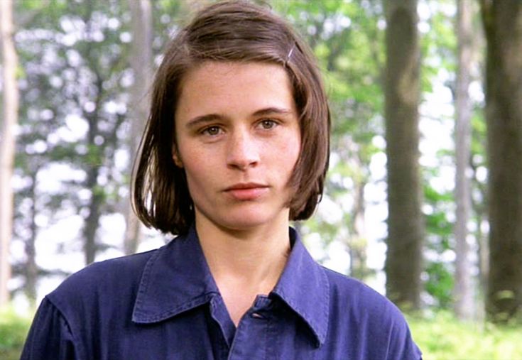 a young man in a blue shirt standing in front of some trees and looking at the camera