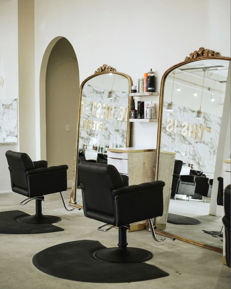 Two black salon styling chairs in front of large gold mirrors with a sand-colored styling station. Salon Chair Design, Two Chair Salon Suite, Salon Suite Decor Black And Gold, Black Hair Salon Decor, Boujee Hair Salon, Hair Salon Mirror, Salon Suite Grand Opening Ideas, Upscale Salon Decor Ideas, Salon Interior Design Black