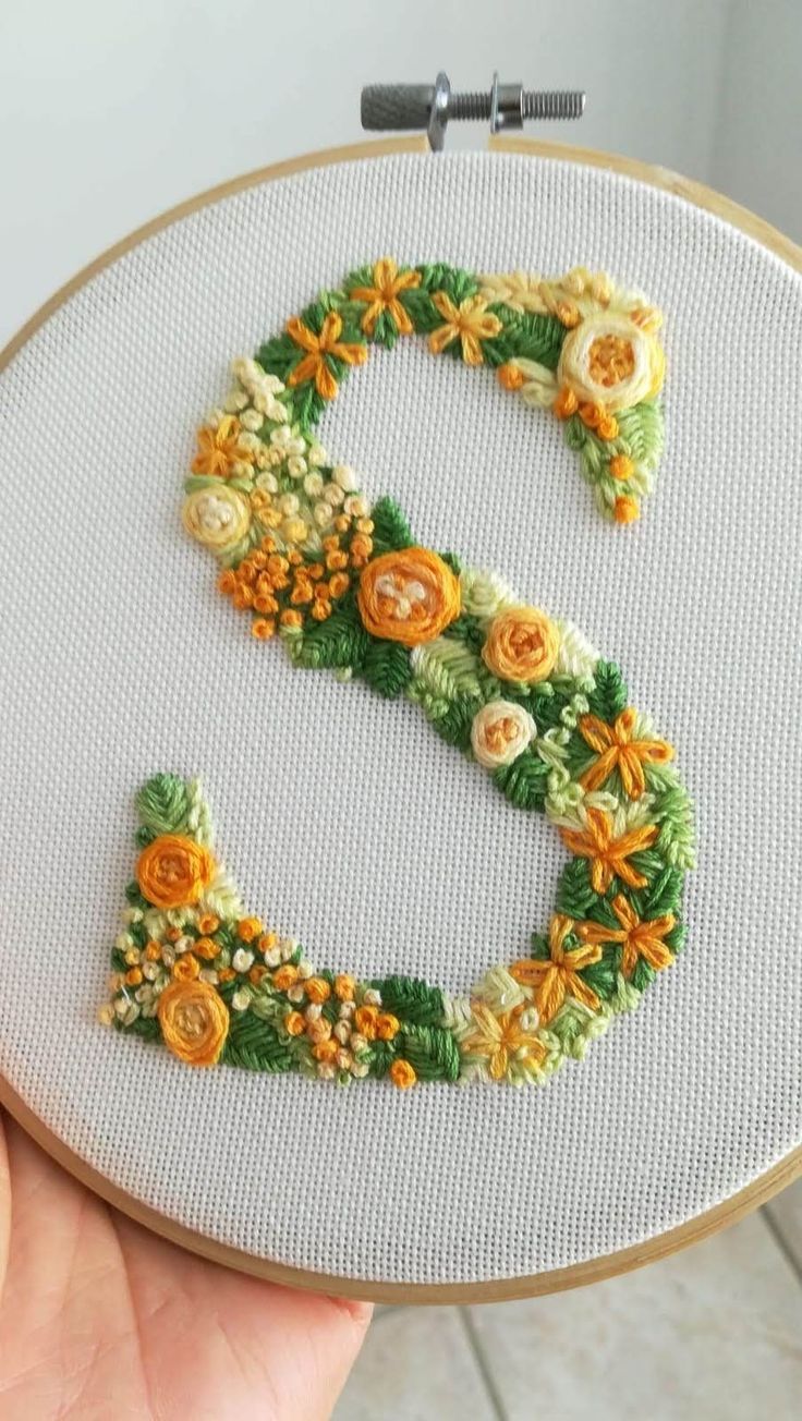 someone is holding up a cross - stitch letter e with flowers on it in front of a white wall