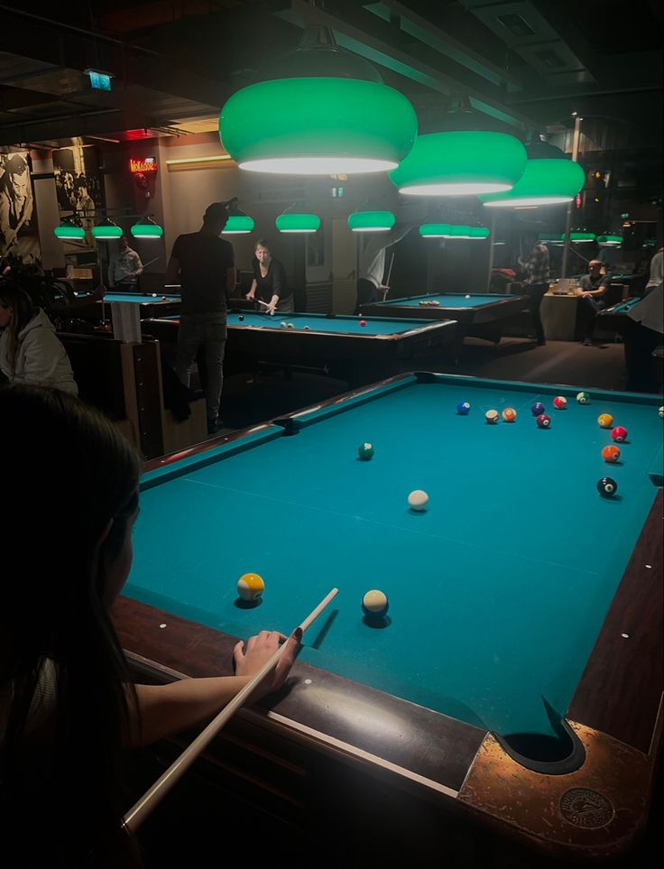 people playing pool in a dark room with green lights on the ceiling and bill tables