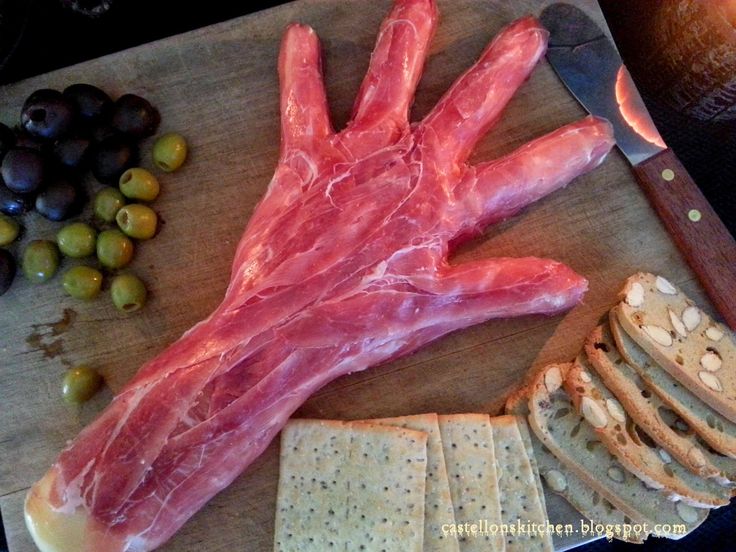 a wooden cutting board topped with meat and crackers next to olives, bread