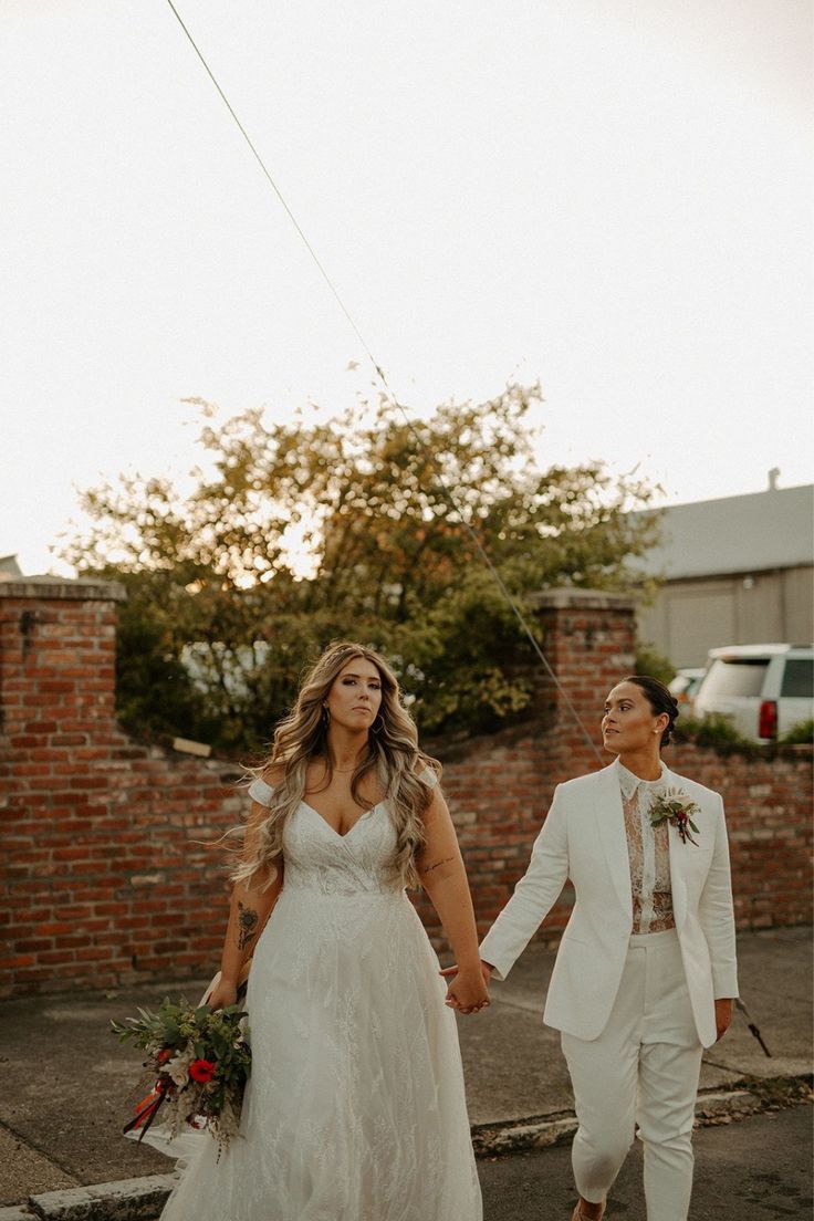 a bride and groom holding hands while walking down the street in front of a brick wall