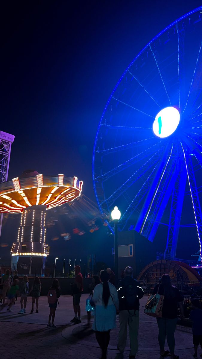 people are walking around in front of an amusement park ride at night with bright lights