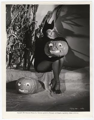 black and white photograph of woman in costume with pumpkin mask sitting next to cat statue