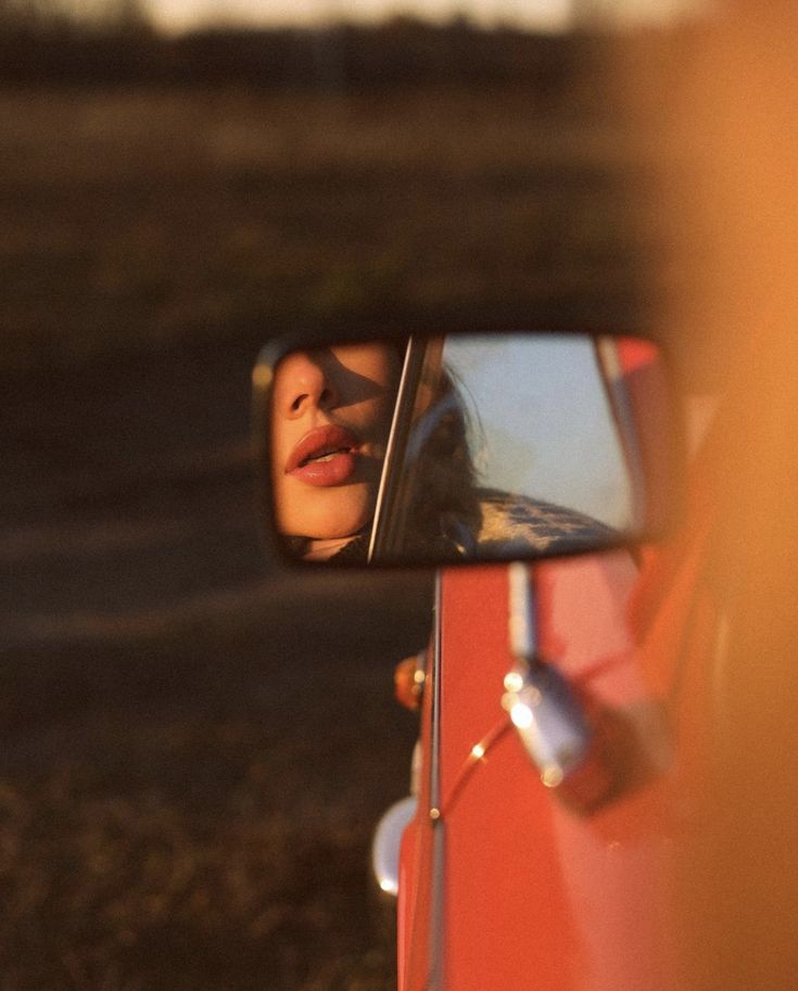 a woman's reflection in the side view mirror of a red car as she looks at her face