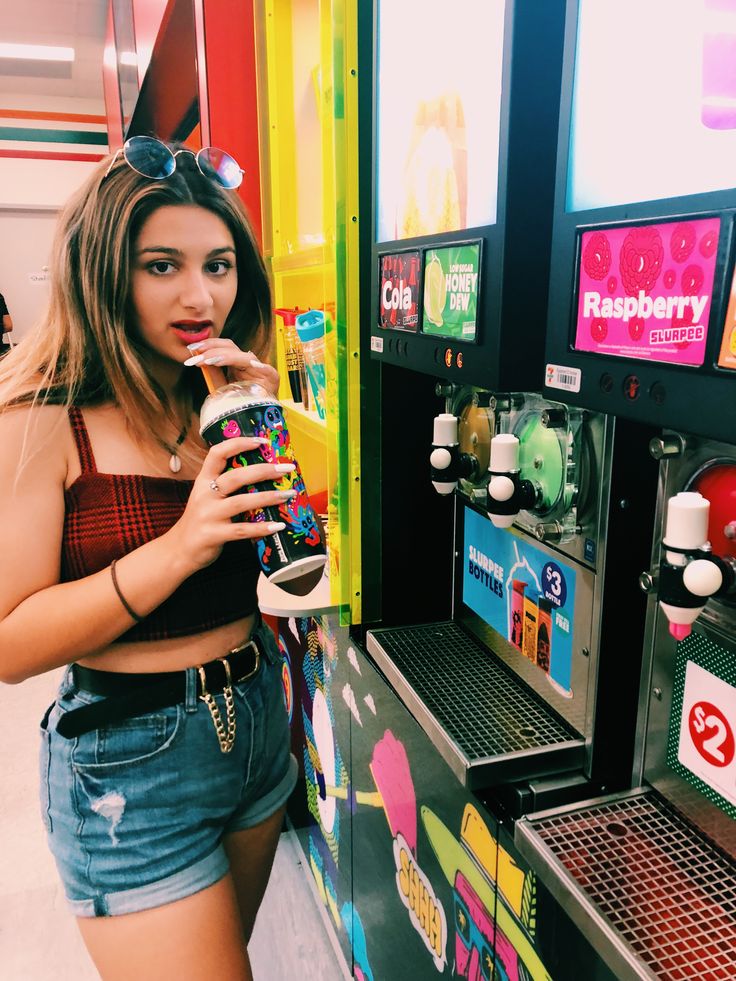 a woman standing in front of a vending machine drinking from a drink cup and holding a soda