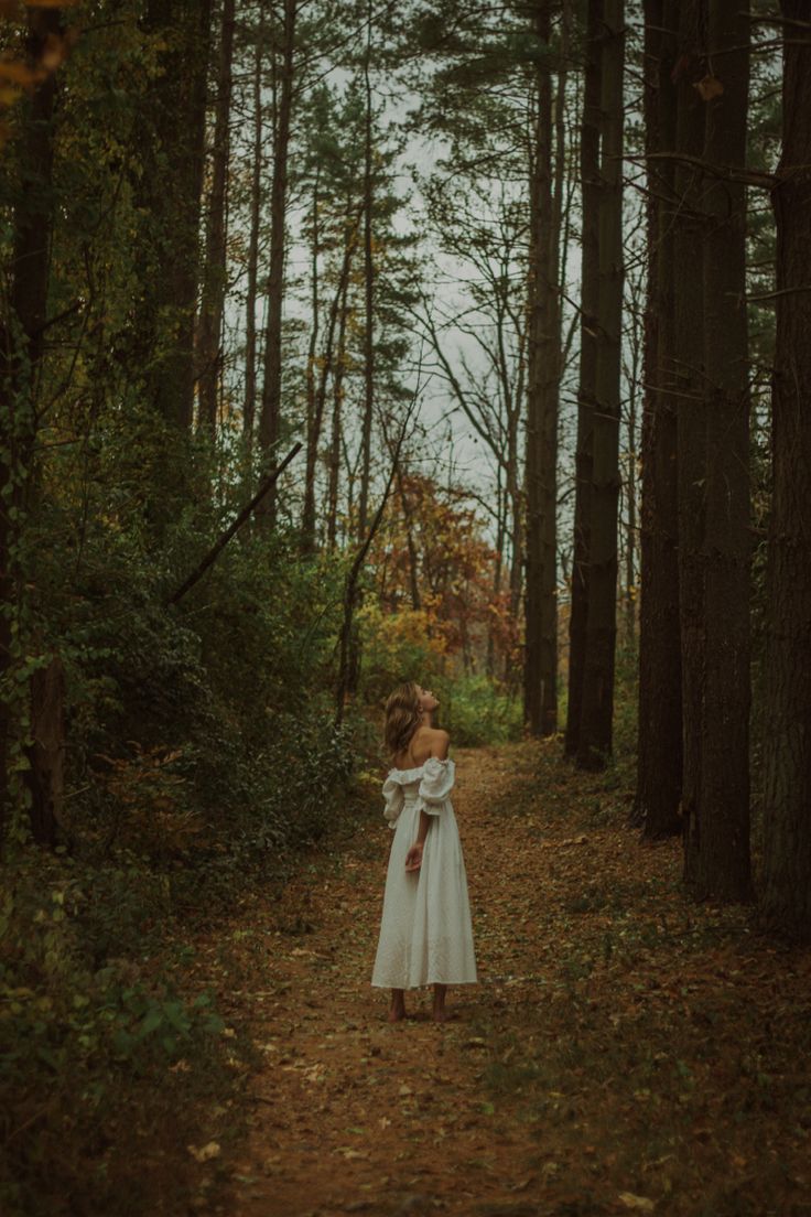 a woman in a white dress walking down a path through the woods with trees on both sides