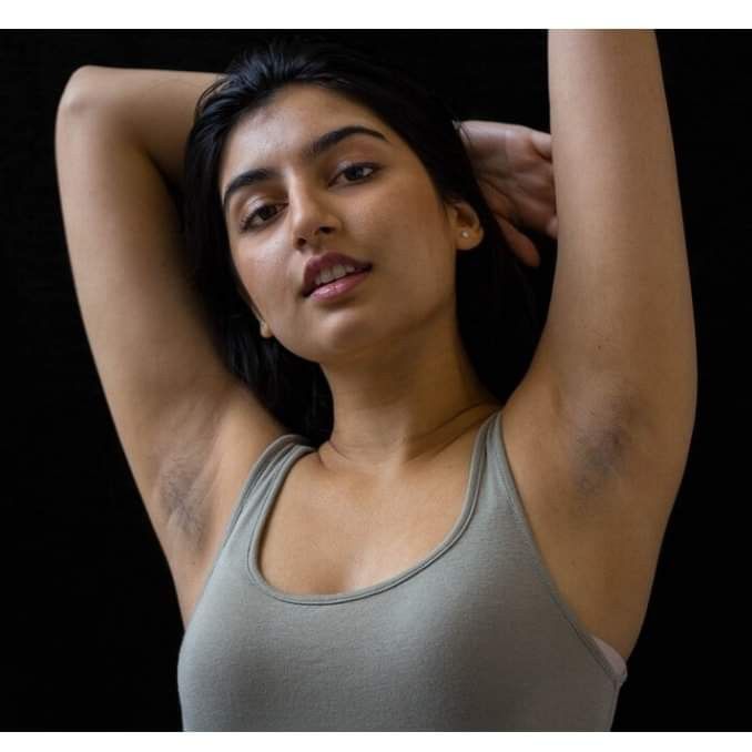 a woman in grey tank top holding her arm behind her head and looking at the camera