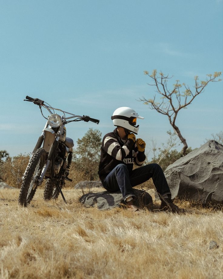 a man sitting on the ground next to his motorcycle and looking at something off in the distance