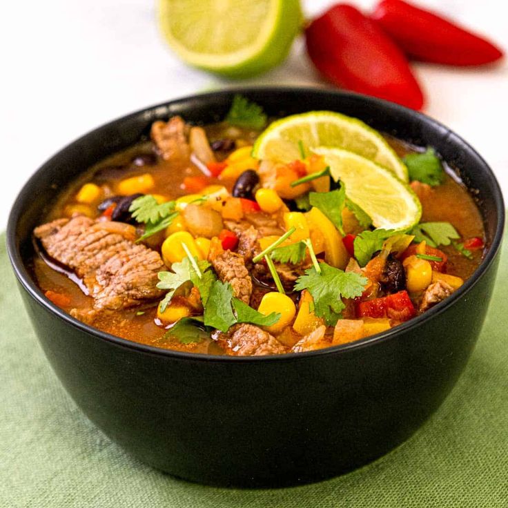 a black bowl filled with meat and vegetables next to sliced limes on a table