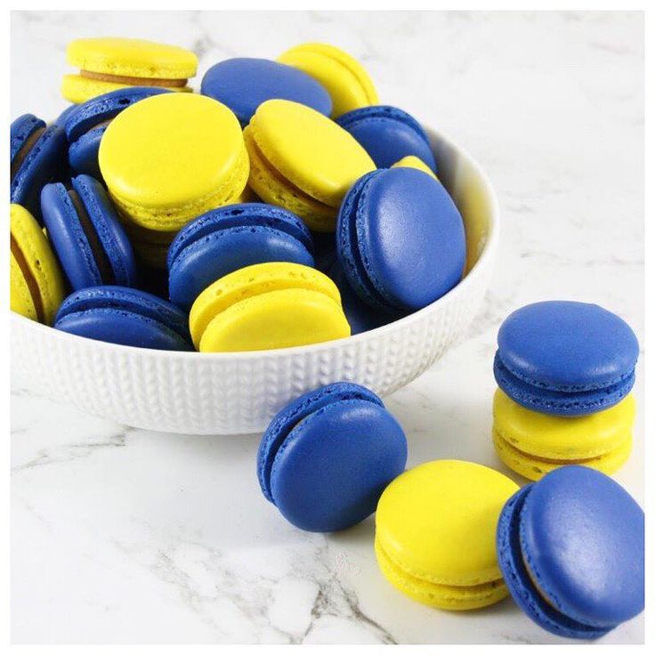 blue and yellow macaroons in a white bowl on a marble countertop next to other macaroons