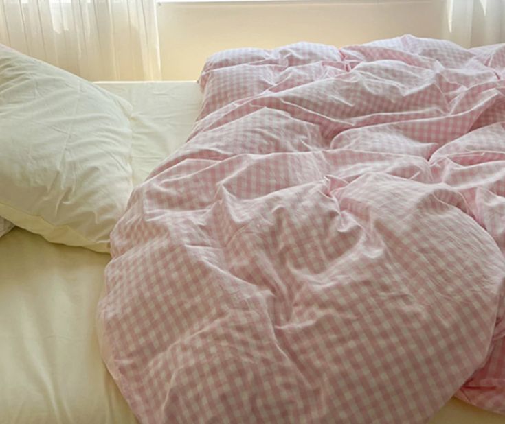 an unmade bed with pink gingham comforter and pillows in front of a window