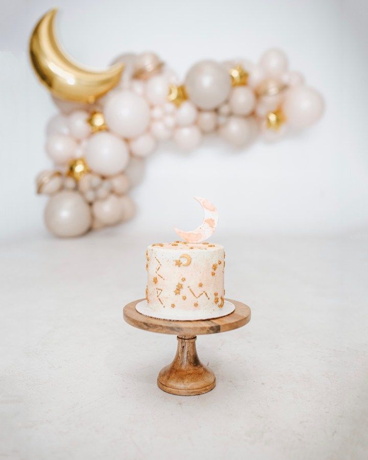 a white cake sitting on top of a wooden stand next to balloons and a crescent