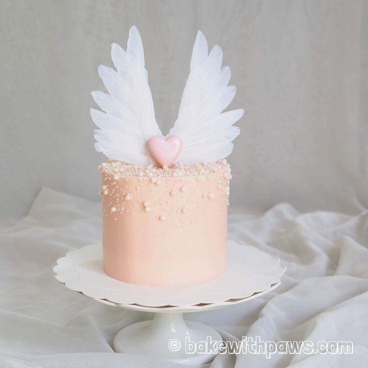 a pink cake with white angel wings and a heart decoration on the top is sitting on a plate