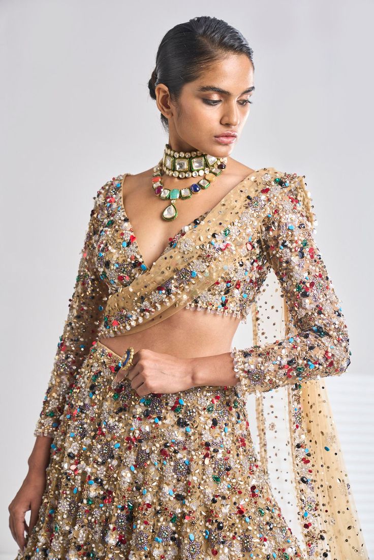 This antique gold net lehenga set has a dense sprinkling of all jewel tones of embroidery material - emerald, ruby, turquoise, gold, silver, pearl etc. It comes with a full sleeves blouse which has delicate tasselling at the sleeve hem and at the waist. The dupatta has four-sided border and dense sprinkling.From Seema Gujral’s Samode collectionDELIVERY TIMEPlease wait 8-12 weeks for your outfit to arrive.FABRIC DETAILSNetProfessional cleaning only. Elegant Pre-draped Kundan Saree For Party, Gold Hand Embellished Sharara For Designer Wear, Gold Embellished Choli, Embellished Gold Choli, Glamorous Gold Hand Embellished Sharara, Glamorous Gold Hand-embellished Sharara, Gold Hand-embellished Sets For Reception, Gold Hand Embellished Sets For Reception, Festive Gold Embellished Choli