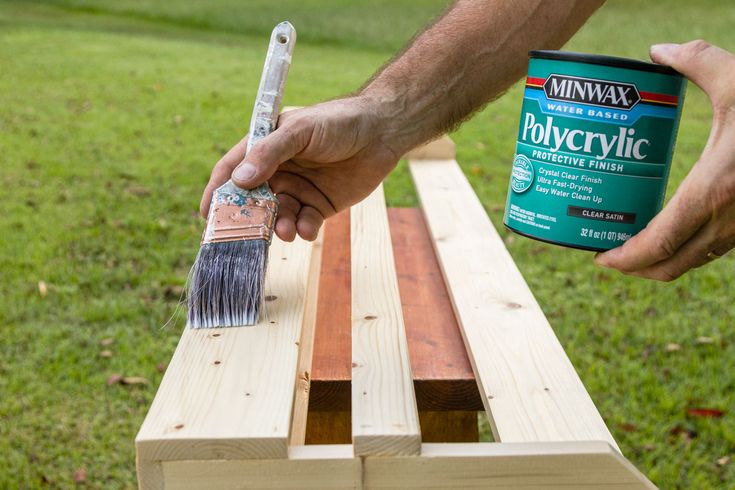 a person holding a paintbrush and painting a wooden bench in front of some grass