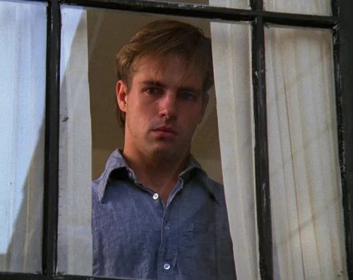 a young man looking out the window with his hand on his hip and wearing a blue shirt