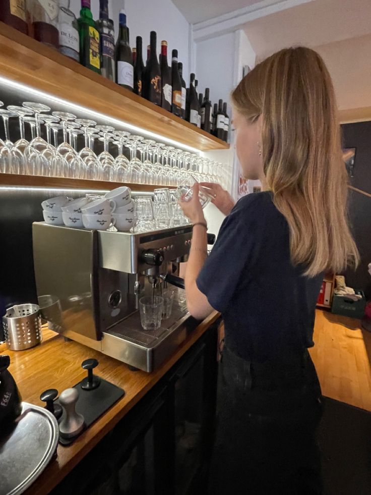 a woman standing in front of a counter filled with wine glasses and cups next to an espresso machine