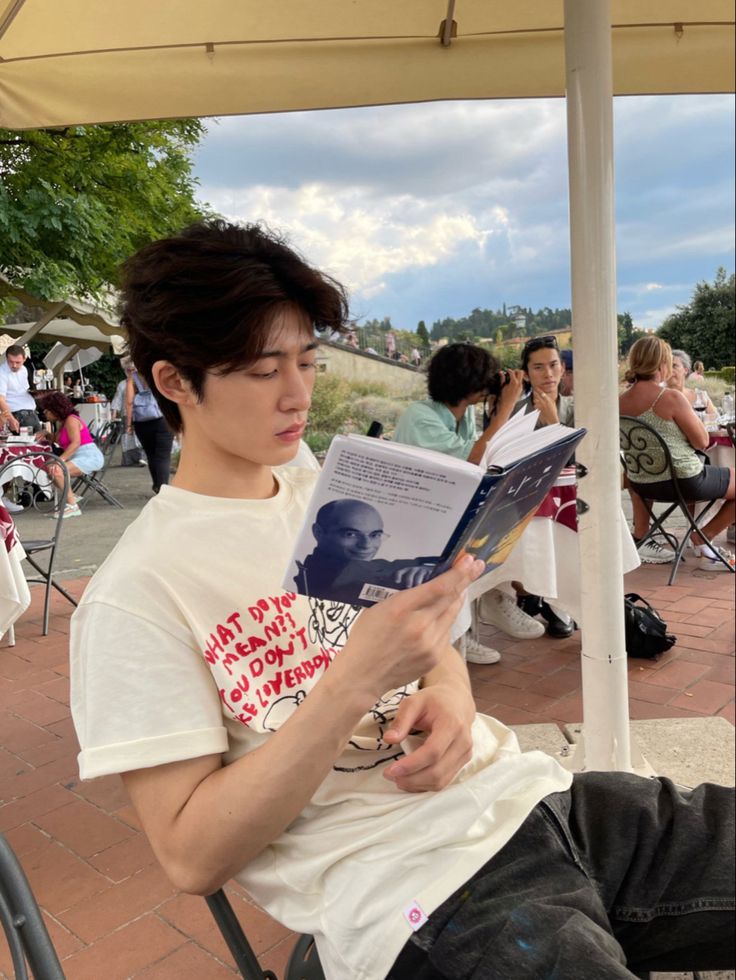 a young man sitting under an umbrella reading a book while others sit in chairs around him