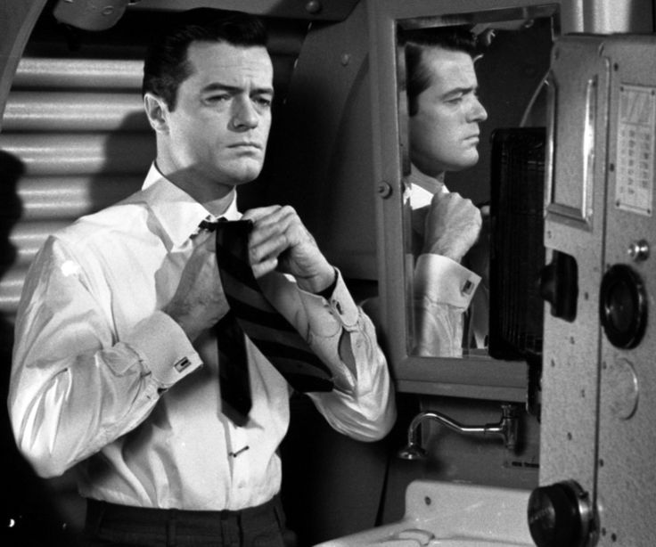 a man adjusting his tie in front of a mirror with another man looking at him