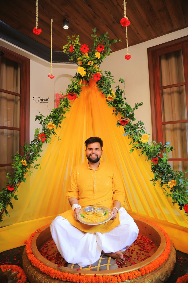 a man sitting in front of a cake on top of a table with flowers hanging from the ceiling