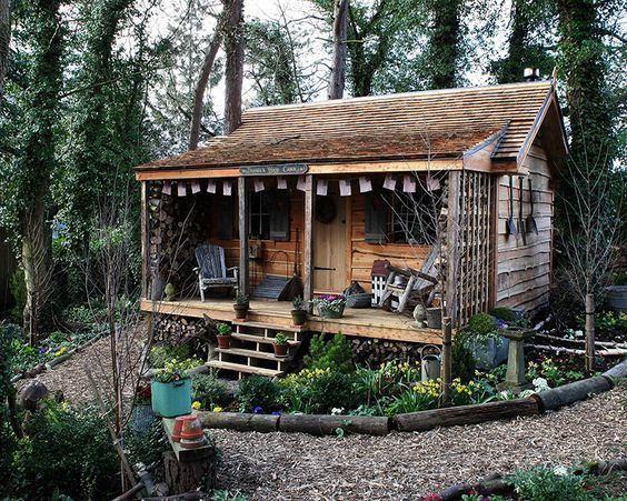 a small wooden cabin in the middle of some trees and bushes with chairs on it