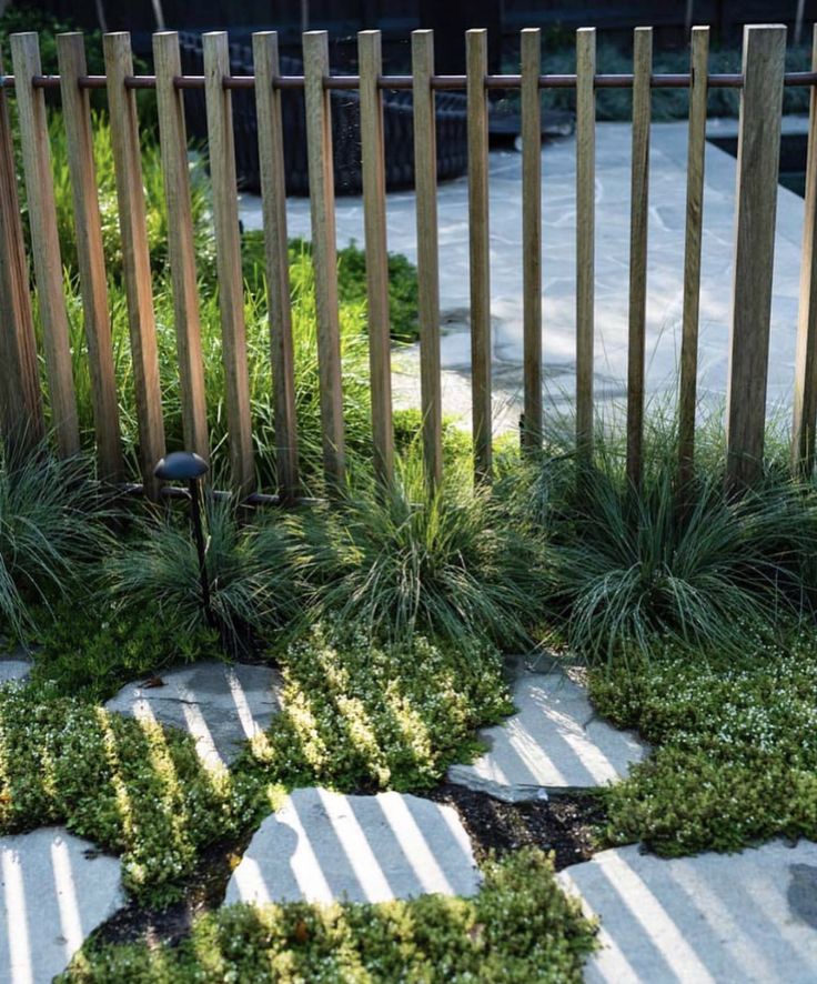 a small garden with grass and rocks in front of a wooden fence on the sidewalk