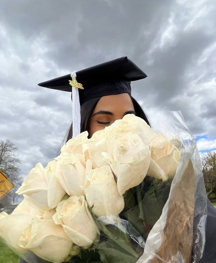 a woman wearing a graduation cap and gown holding flowers in front of her face with cloudy skies behind her