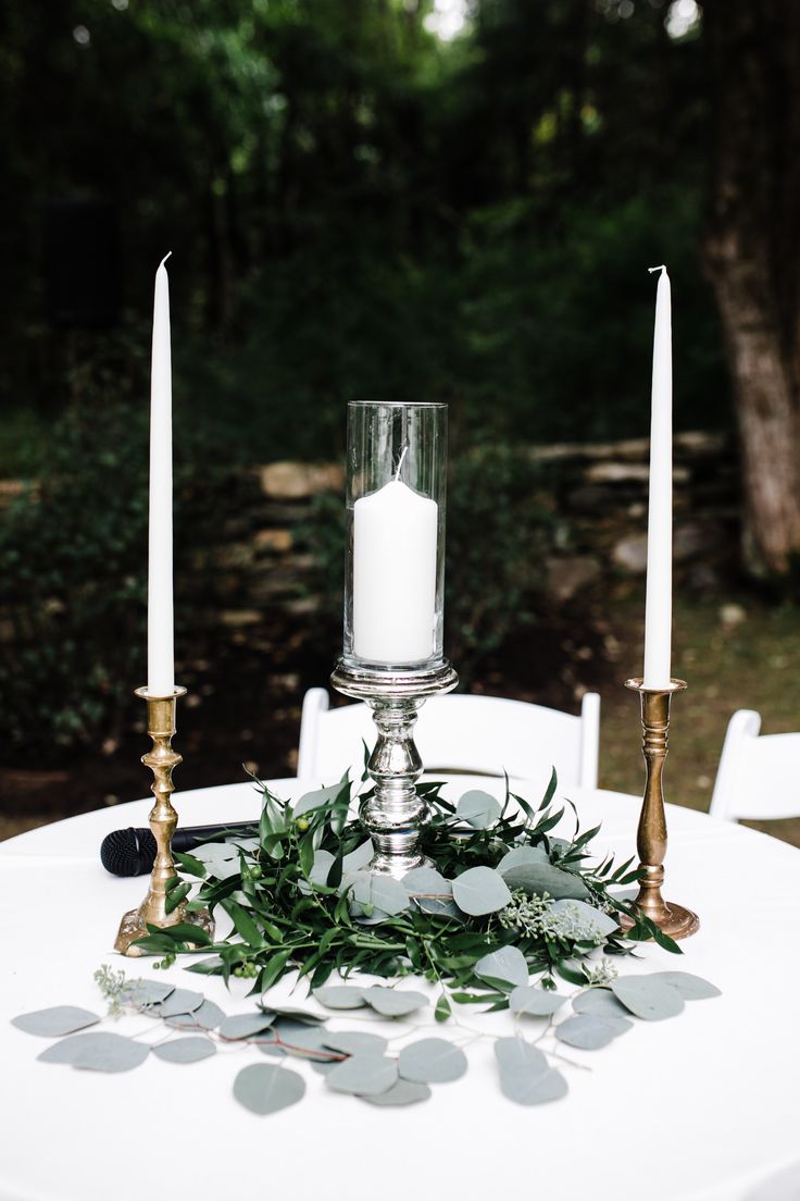 two candles are sitting on top of a white table cloth with greenery around it