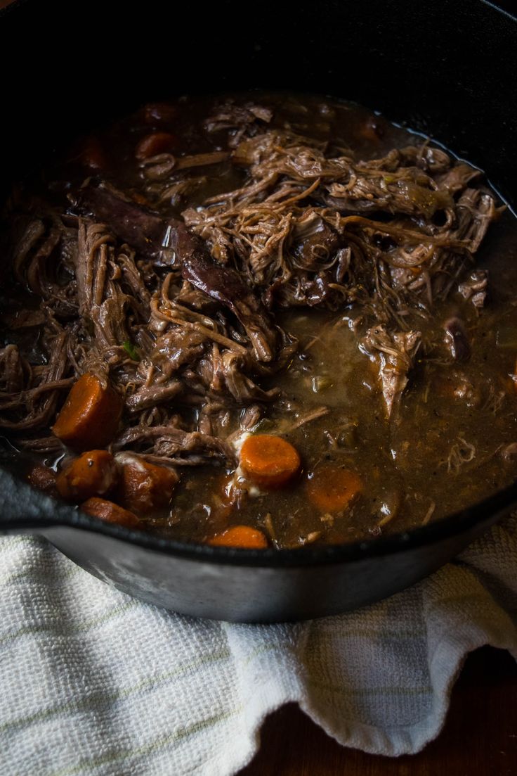 a pot filled with meat and carrots on top of a wooden table next to a white towel